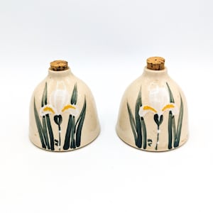 May include: Two ceramic salt and pepper shakers with cork tops. Each shaker features a white iris flower with yellow accents painted on a beige background. The flowers are surrounded by green leaves.