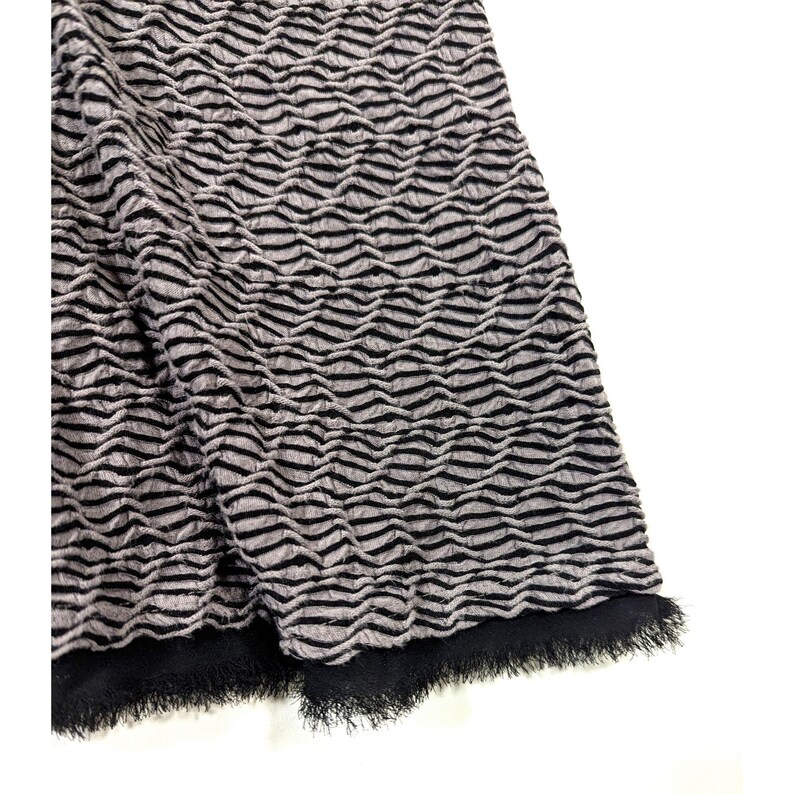 May include: Close-up of a textured fabric with a wave-like pattern in shades of gray and black. The fabric has a fringed edge in black. The material appears to be soft and pliable, suitable for clothing or home decor.