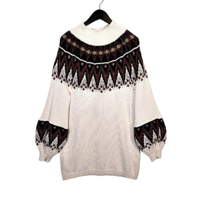 Puede incluir: Su&eacute;ter color crema con cuello alto y mangas largas. La parte superior del pecho y las mangas presentan un estampado geom&eacute;trico en marr&oacute;n y negro. El su&eacute;ter est&aacute; colgado de una percha negra sobre un fondo blanco.
