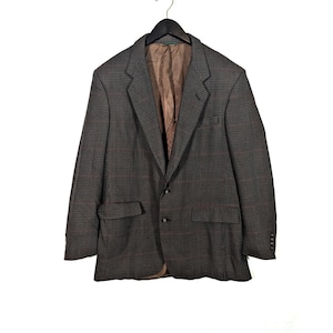 Può includere: Un blazer a quadri grigio con fodera marrone, appeso a una gruccia nera. Il blazer ha una classica chiusura a due bottoni, due tasche anteriori e un revers a lancia. Il motivo a quadri include linee rosse e nere.