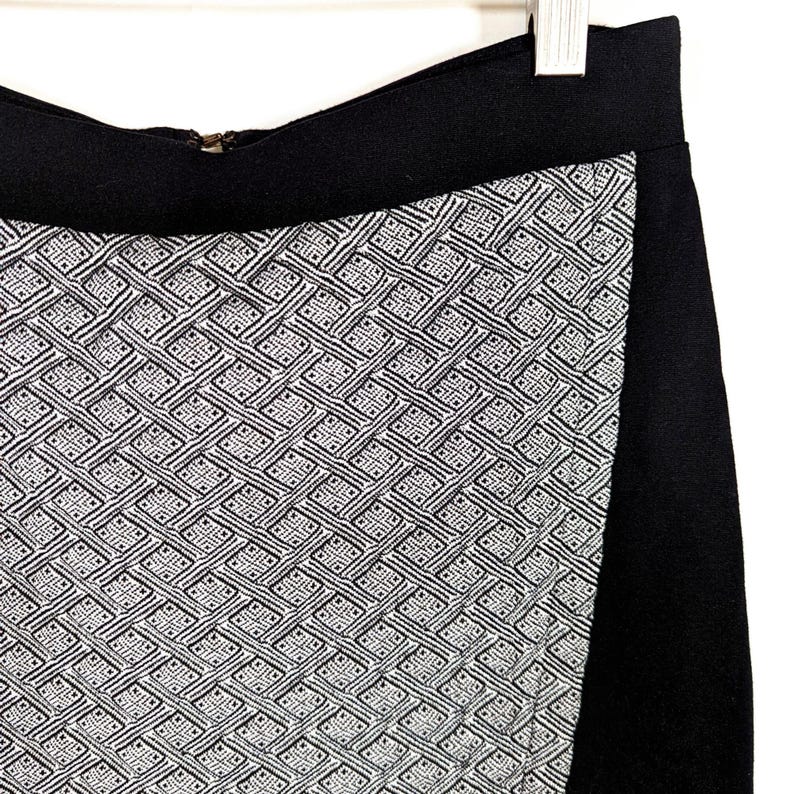 May include: A black skirt with a geometric pattern in white and black. The skirt has a wide black waistband and a side panel of solid black fabric. The geometric pattern is a repeating design of interlocking shapes.
