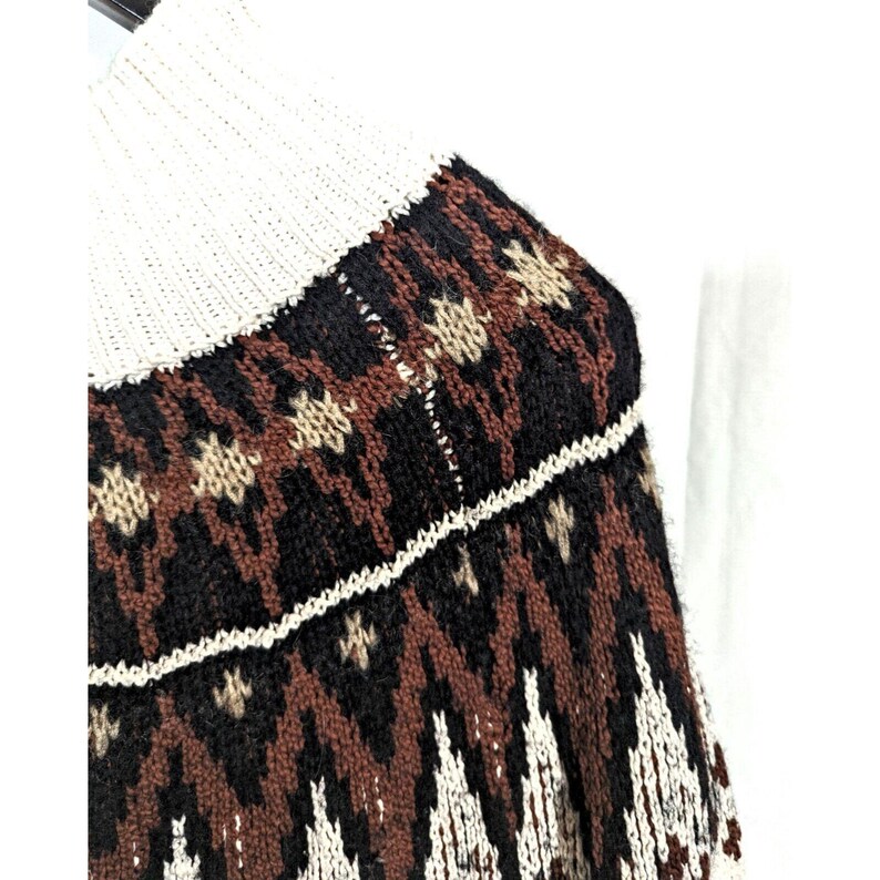 Puede incluir: Primer plano de un su&eacute;ter de punto con cuello alto color crema. El su&eacute;ter presenta un patr&oacute;n geom&eacute;trico en negro, marr&oacute;n y crema. El dise&ntilde;o incluye formas de diamante y estrella.