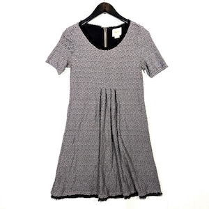 May include: A short-sleeved dress with a black and gray textured pattern. The dress has a black neckline and a zipper closure. The skirt is pleated, and the hemline is trimmed with black fringe. The dress is hanging on a black hanger.
