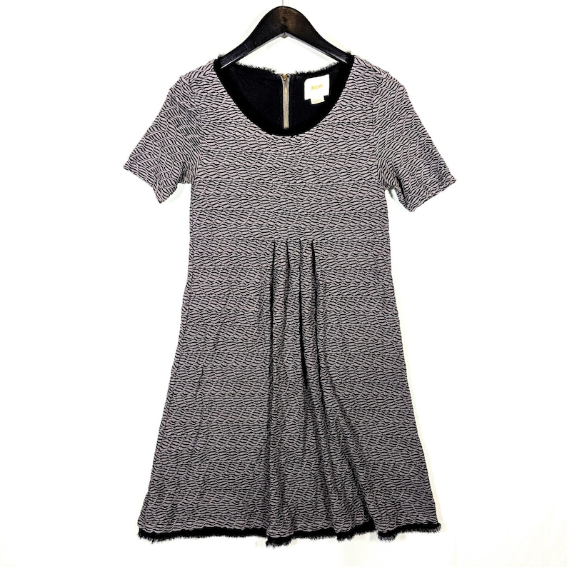 May include: A short-sleeved dress with a black and gray textured pattern. The dress has a black neckline and a zipper closure. The skirt is pleated, and the hemline is trimmed with black fringe. The dress is hanging on a black hanger.