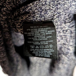 K&ouml;nnte beinhalten: Nahaufnahme eines schwarzen Kleidungsetiketts mit wei&szlig;em Text. Das Etikett listet die Stoffzusammensetzung als Baumwolle, Merinowolle und Rayon auf. Das Etikett enth&auml;lt auch Waschanweisungen und den Satz "Made in China."