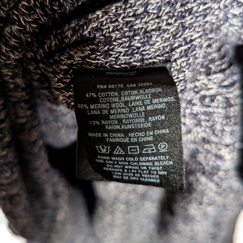 K&ouml;nnte beinhalten: Nahaufnahme eines schwarzen Kleidungsetiketts mit wei&szlig;em Text. Das Etikett listet die Stoffzusammensetzung als Baumwolle, Merinowolle und Rayon auf. Das Etikett enth&auml;lt auch Waschanweisungen und den Satz "Made in China."