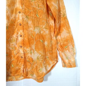 Puede incluir: Camisa naranja con botones, mangas largas y un bolsillo. La camisa presenta dise&ntilde;os florales bordados en verde y blanco. La tela tiene un efecto tie-dye con parches naranja claro y blanco.