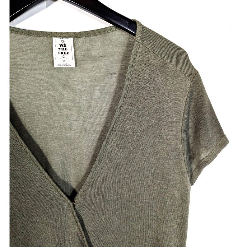 May include: Olive green wrap top with short sleeves. The top has a V-neck design and a tag that reads "WE THE FREE". The top is hanging on a black hanger.