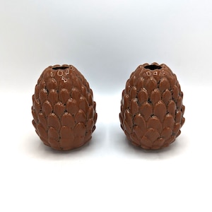 Könnte beinhalten: Zwei braune Keramikvasen in Form von Tannenzapfen. Die Vasen haben eine strukturierte Oberfläche und sind dunkelbraun.