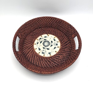 May include: A round woven wicker tray with two handles and a decorative ceramic tile in the centre. The tile is white with a blue floral design.