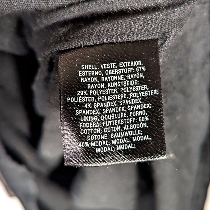 May include: A close-up of a black fabric tag with white text. The tag lists the materials of a garment, including rayon, polyester, spandex, cotton, and modal. The tag also includes the word "shell" and "lining".