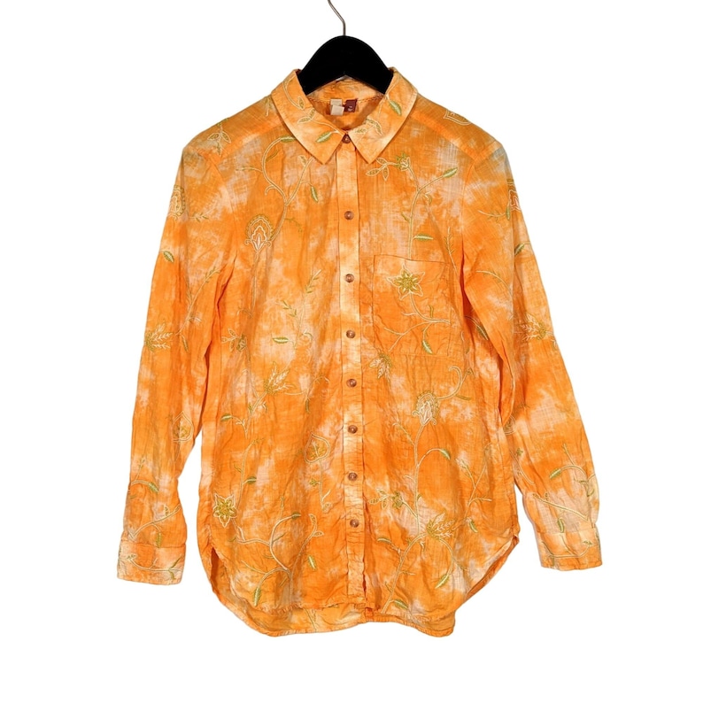 Puede incluir: Camisa naranja con botones y mangas largas, con dise&ntilde;o de bordado floral. La camisa est&aacute; colgada en una percha negra. La tela tiene un efecto tie-dye con acentos blancos y verdes.