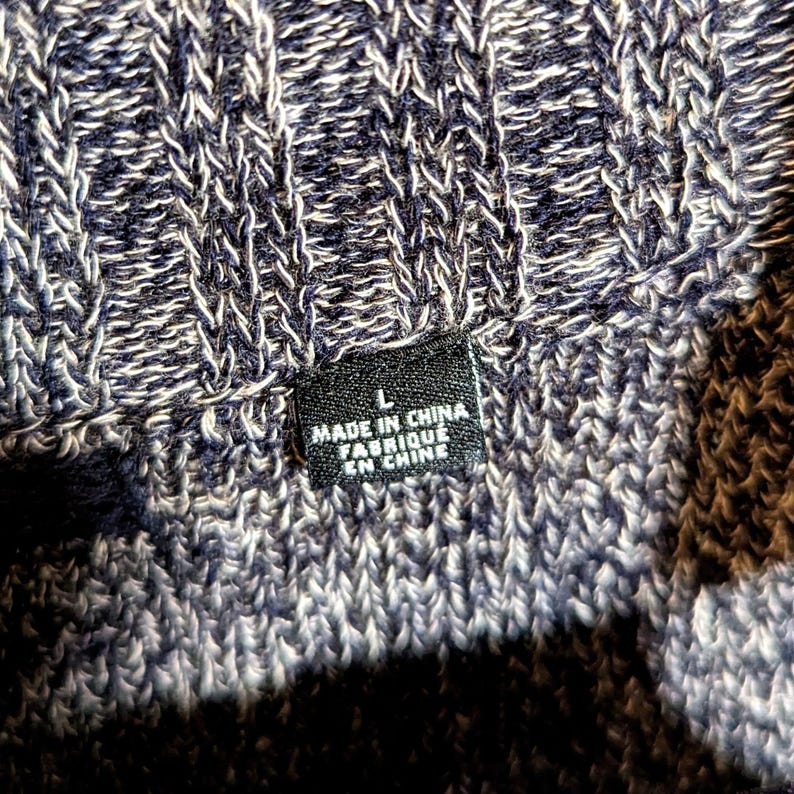 K&ouml;nnte beinhalten: Nahaufnahme eines strukturierten Strickstoffs in Marineblau und Wei&szlig;. Ein schwarzes Etikett mit dem Text "L MADE IN CHINA FABRIQUE EN CHINE" ist sichtbar. Das Strickware hat ein Rippenmuster.