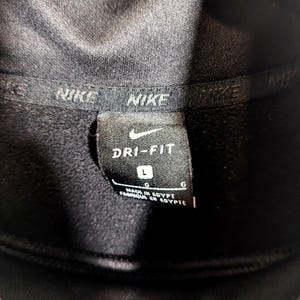 May include: Close-up of a black Nike Dri-FIT athletic garment tag. The tag features the Nike swoosh logo, the words "DRI-FIT", and the size "L". The tag also indicates the garment was made in Egypt.