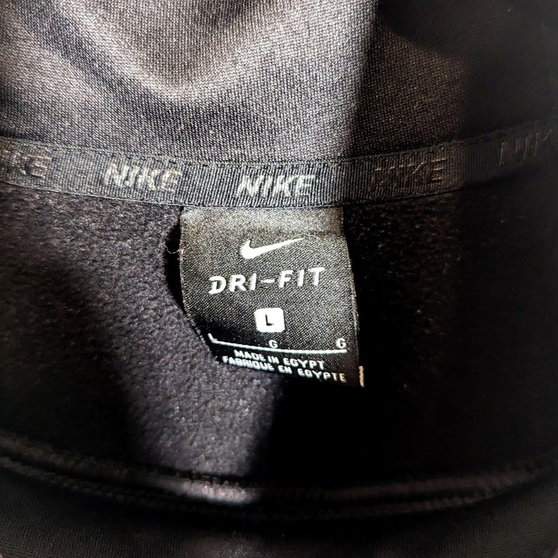 May include: Close-up of a black Nike Dri-FIT athletic garment tag. The tag features the Nike swoosh logo, the words "DRI-FIT", and the size "L". The tag also indicates the garment was made in Egypt.