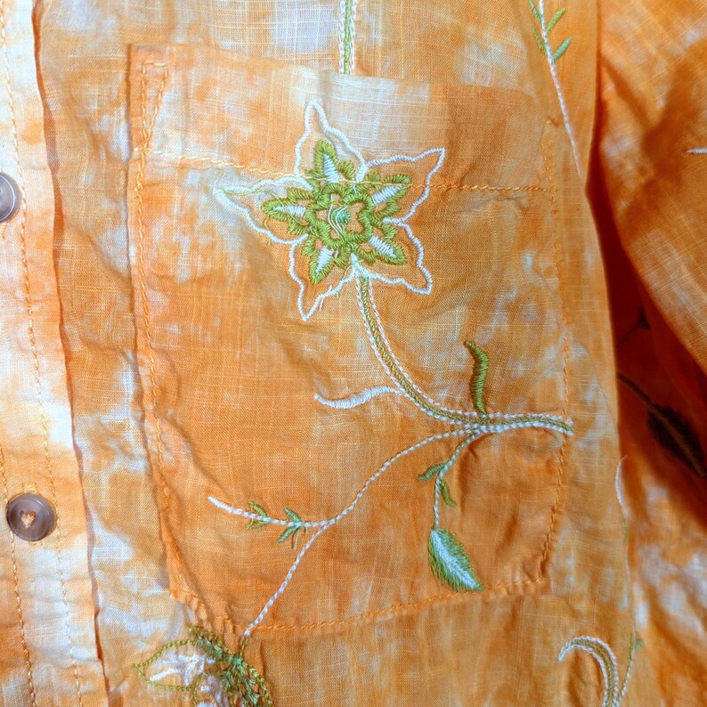 Puede incluir: Primer plano de una camisa naranja con botones y bordado floral en el bolsillo. El bordado presenta una flor verde y blanca con un tallo y hojas. La camisa tiene un efecto tie-dye.
