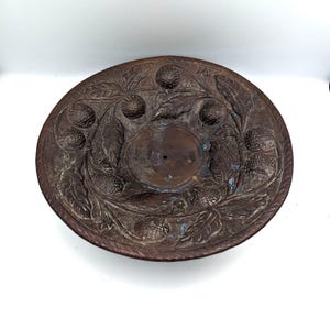 May include: A round, copper-colored metal dish with an embossed design of leaves and berries. The dish has a raised edge and a small hole in the center.