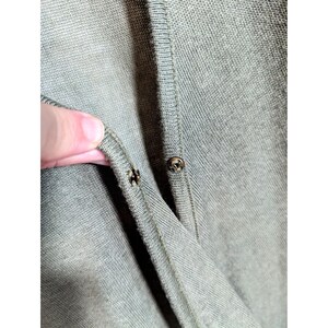May include: Close-up of a sage green cardigan with two metal snap closures. The fabric is a fine knit, and the buttons are small and round. The cardigan is partially unbuttoned, revealing the inside of the garment.