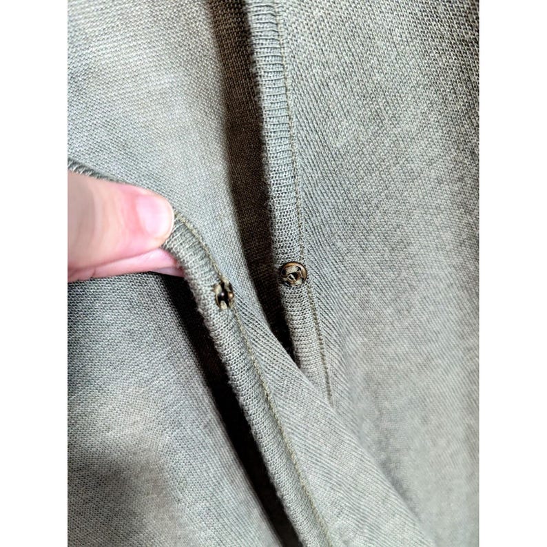May include: Close-up of a sage green cardigan with two metal snap closures. The fabric is a fine knit, and the buttons are small and round. The cardigan is partially unbuttoned, revealing the inside of the garment.