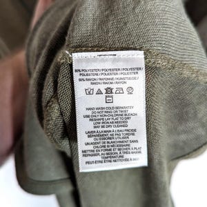 May include: Close-up of a green garment label with care instructions. The label includes text detailing washing, drying, and ironing instructions. The fabric composition is listed as 50% polyester and 50% rayon.