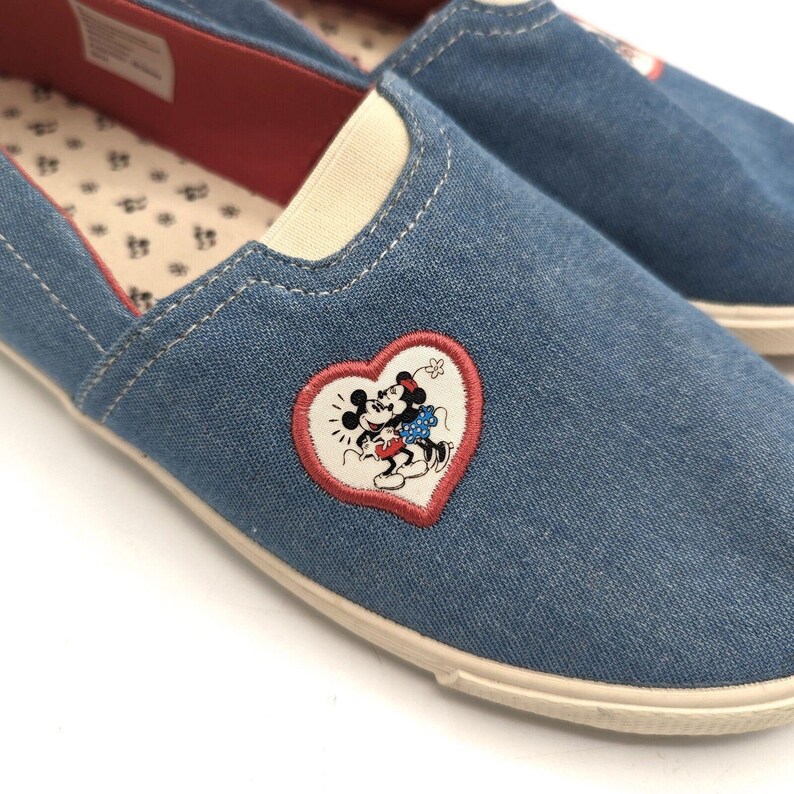 May include: Blue denim slip-on shoes with a heart-shaped patch featuring a Minnie and Mickey Mouse design. The shoes have a red interior and a cream sole. A casual style of footwear.