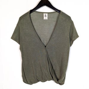 May include: Olive green wrap-style top with short sleeves. The top features a deep V-neckline and a knot detail at the waist. A small tag with the text "WE THE FREE" is visible near the neckline. The top is hanging on a black hanger.