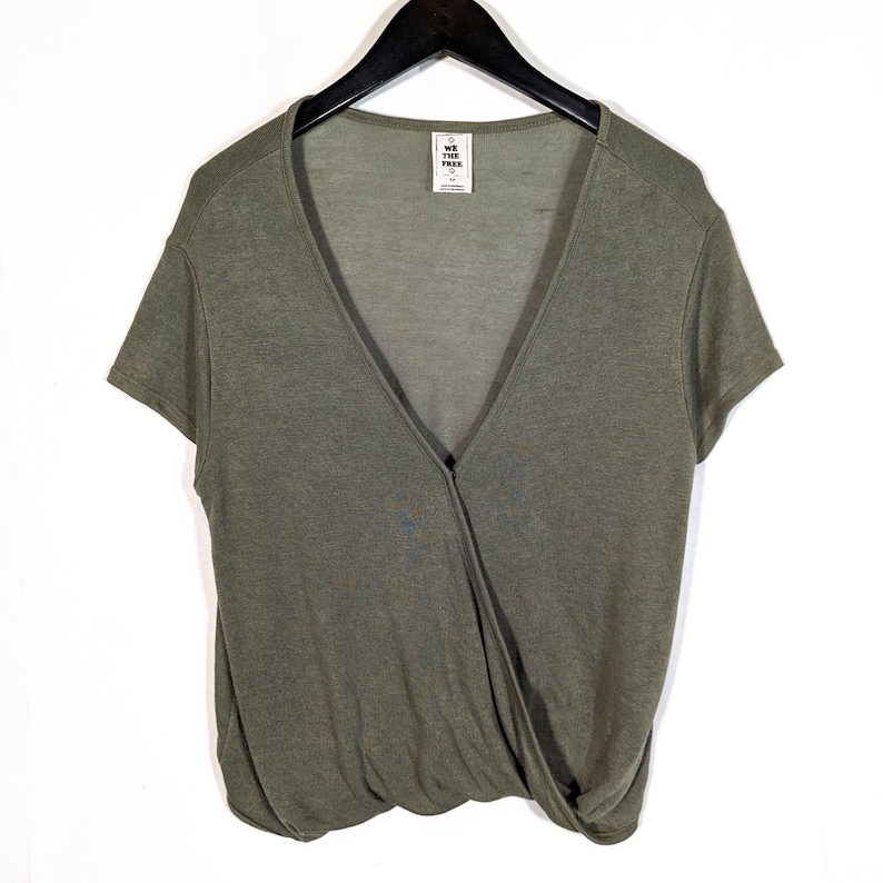 May include: Olive green wrap-style top with short sleeves. The top features a deep V-neckline and a knot detail at the waist. A small tag with the text "WE THE FREE" is visible near the neckline. The top is hanging on a black hanger.
