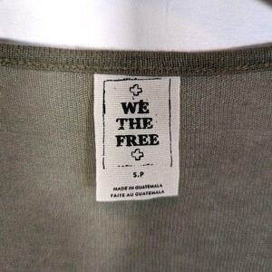 May include: Close-up of a green garment with a tag that reads "WE THE FREE" in black text. The tag also includes the text "MADE IN GUATEMALA" and "FAITE AU GUATEMALA". The garment has a ribbed neckline.