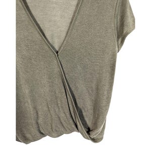 May include: A sage green wrap-style top with a V-neckline and short sleeves. The fabric appears to be a soft knit material. The top has a draped design that gathers at the waist.