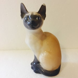 Puede incluir: Una figurita de porcelana de un gato siamés sentado con la cola enroscada alrededor de sus patas. El gato es blanco con marcas marrones en la cara, las orejas y la cola. La figurita está esmaltada y tiene un acabado brillante.