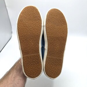 May include: A pair of blue canvas trainers with white rubber soles. The soles are brown with a textured pattern. The trainers are held up against a white background.