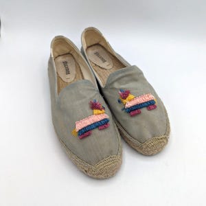Puede incluir: Un par de zapatos sin cordones de alpargata grises con diseños de piñata bordados. Las piñatas presentan detalles en rosa, amarillo, azul y rojo. Los zapatos tienen una suela de yute tejida y la marca "Soludos" es visible en la plantilla.
