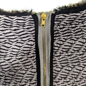 May include: Close-up of a textured fabric with a repeating wave pattern in gray and black. A gold-colored zipper runs down the center, framed by a gray and black trim. The top edge has a black fringe.