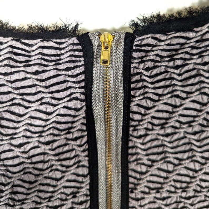 May include: Close-up of a textured fabric with a repeating wave pattern in gray and black. A gold-colored zipper runs down the center, framed by a gray and black trim. The top edge has a black fringe.