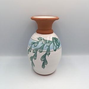 Può includere: Un vaso in ceramica con base bianca, decorato con una striscia azzurra e un motivo a foglie verdi. La parte superiore e il collo del vaso sono color terracotta. Il vaso ha un corpo arrotondato e un bordo svasato.