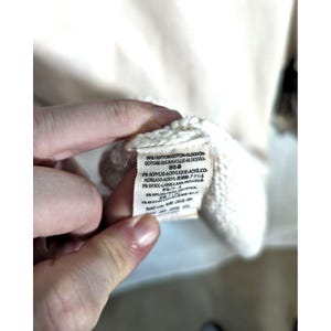 Puede incluir: Primer plano de una etiqueta de tela color crema con texto en varios idiomas. La etiqueta est&aacute; sujeta por una mano y est&aacute; adherida a un su&eacute;ter color crema. La etiqueta enumera la composici&oacute;n de la tela, incluyendo algod&oacute;n, acr&iacute;lico, lana y nailon.