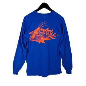May include: A long-sleeved blue shirt with an orange graphic of a fish and the words "Salt Life". The graphic is on the back of the shirt. The shirt is hanging on a black hanger.