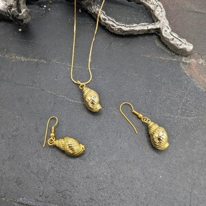 May include: A gold necklace and earrings set featuring a seashell design. The necklace has a delicate chain and a pendant shaped like a seashell. The earrings are also seashell shaped and dangle from small gold hooks.