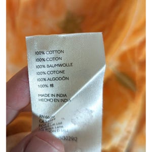 Puede incluir: Una etiqueta de tela blanca con texto que indica 100% algod&oacute;n en varios idiomas. La etiqueta tambi&eacute;n indica "Made in India" e incluye un c&oacute;digo de producto. El fondo es una tela borrosa naranja y amarilla.