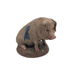 May include: A small, brown pig figurine with a blue ribbon on its back. The pig is sitting on a base that looks like dirt with green leaves.