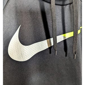 May include: Close-up of a black hooded sweatshirt featuring a large white Nike swoosh logo with a textured pattern. The logo is accented with a neon green stripe. Black drawstrings are visible.