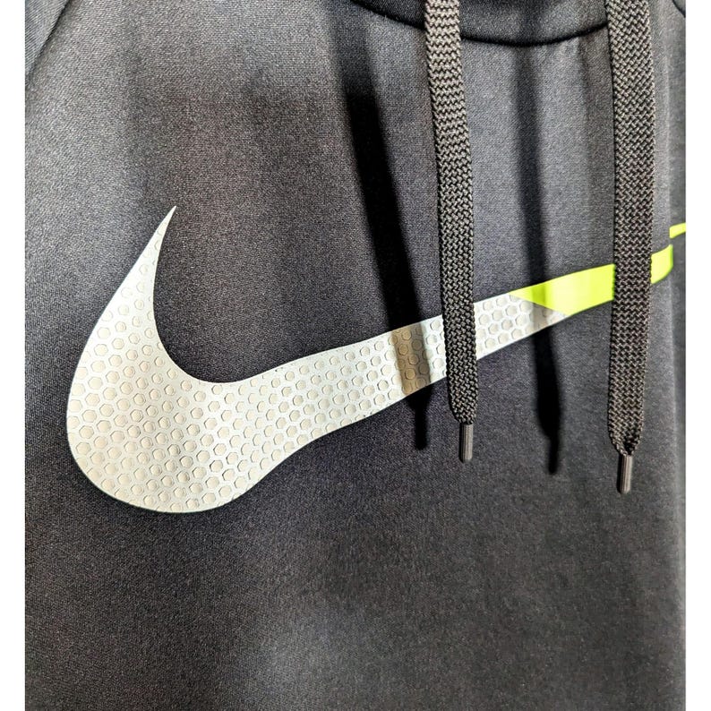 May include: Close-up of a black hooded sweatshirt featuring a large white Nike swoosh logo with a textured pattern. The logo is accented with a neon green stripe. Black drawstrings are visible.