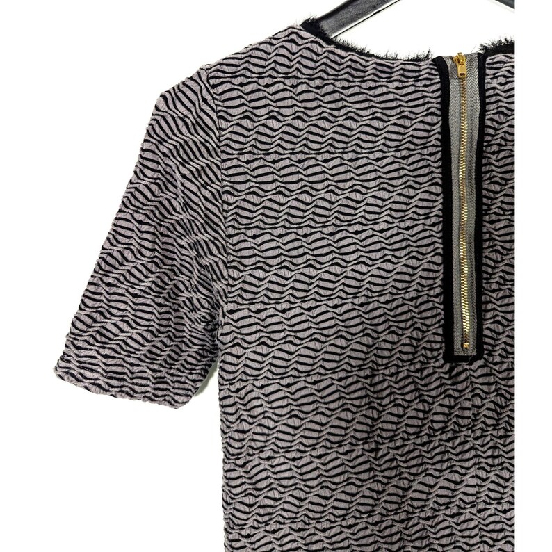 May include: A short-sleeved top with a textured wave pattern in black and light gray. The top features a gold zipper down the back and a black trim around the neckline.