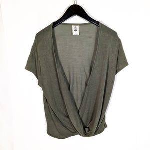 May include: Olive green short-sleeve top with a deep V-neckline and a twisted knot detail at the waist. The top is hanging on a black hanger against a white background. The tag reads "WE THE FREE".