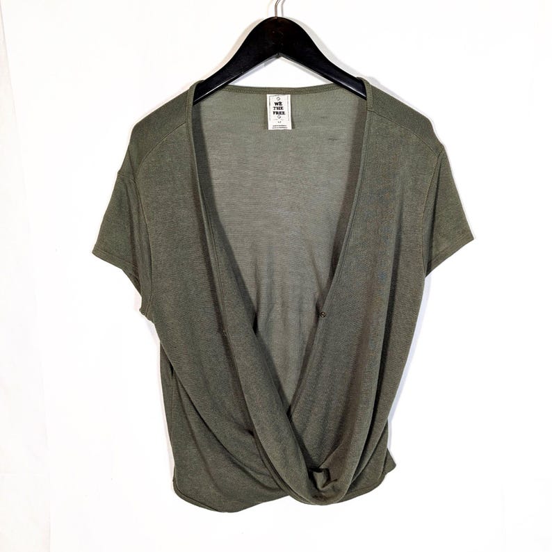 May include: Olive green short-sleeve top with a deep V-neckline and a twisted knot detail at the waist. The top is hanging on a black hanger against a white background. The tag reads "WE THE FREE".