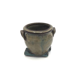 May include: A small, handcrafted ceramic pot with a rounded body and a dark rim. The pot has two small handles and sits on a square base. The exterior features a blend of green and brown hues, creating a unique, earthy aesthetic. Ideal for small plants or decorative use.