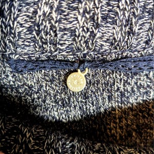 K&ouml;nnte beinhalten: Nahaufnahme eines Strickkleidungsst&uuml;cks in Marineblau und Wei&szlig;. Ein dunkelblauer Besatz mit einem kleinen, runden, goldfarbenen Anh&auml;nger ist angebracht. Der Anh&auml;nger tr&auml;gt die Aufschrift "KNITTED & KNOTTED" in einem Kreis.