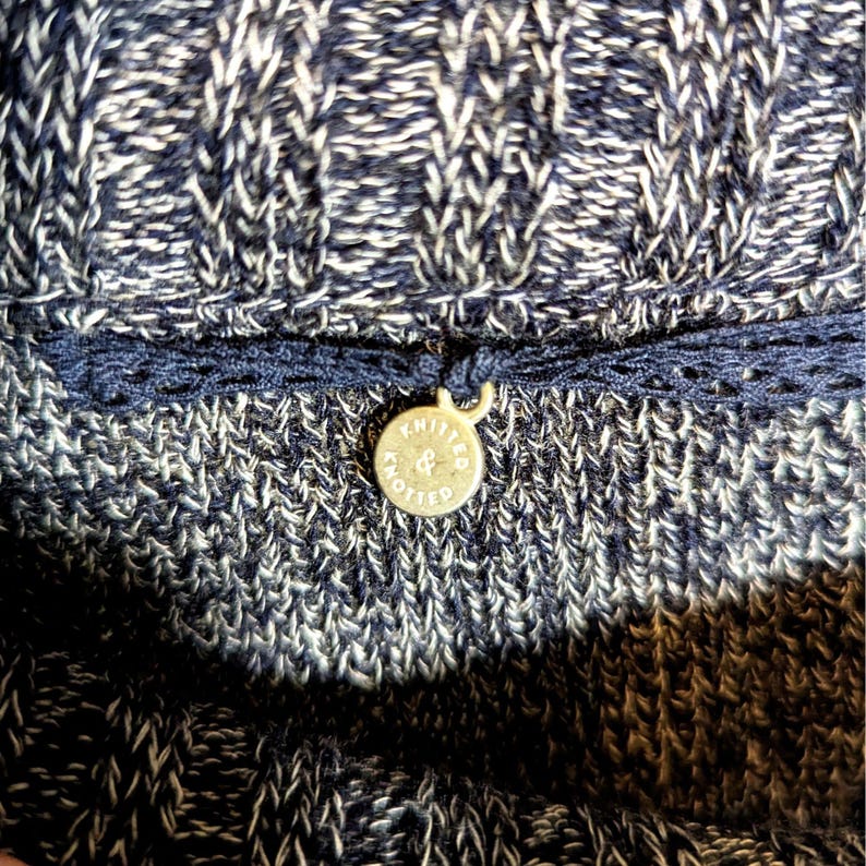 K&ouml;nnte beinhalten: Nahaufnahme eines Strickkleidungsst&uuml;cks in Marineblau und Wei&szlig;. Ein dunkelblauer Besatz mit einem kleinen, runden, goldfarbenen Anh&auml;nger ist angebracht. Der Anh&auml;nger tr&auml;gt die Aufschrift "KNITTED & KNOTTED" in einem Kreis.