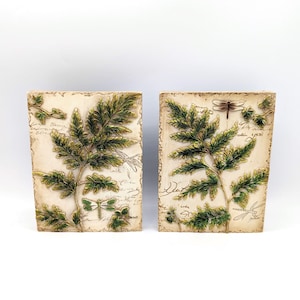 Può includere: Due pannelli decorativi rettangolari dall'estetica vintage. Ogni pannello presenta un rilievo dettagliato di fronde di felci verdi e illustrazioni di libellule su uno sfondo color crema con testo in stile script. I pannelli sono probabilmente per l'arredamento murale.