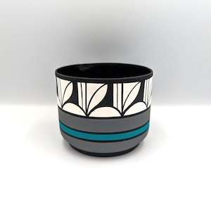 May include: A black and white ceramic bowl with a geometric design. The bowl features a black interior, a gray band with a teal stripe, and a white and black leaf pattern around the top. The bowl is approximately 4 inches in diameter.